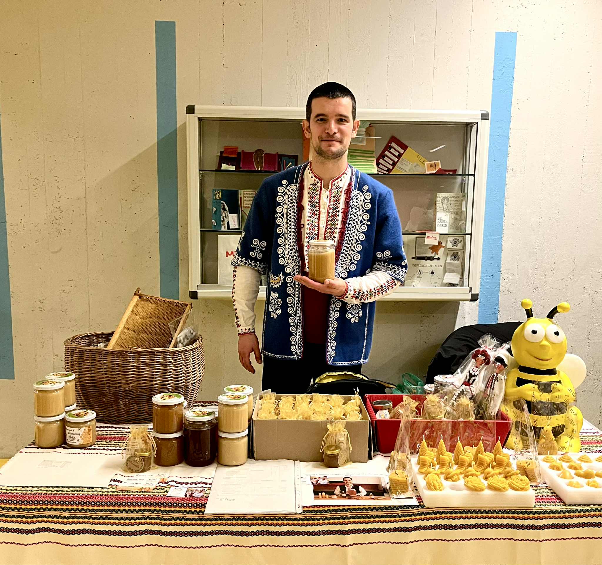Beekeeper in traditional attire with honey jar at a market stall displaying honey, honeycomb, and beeswax products